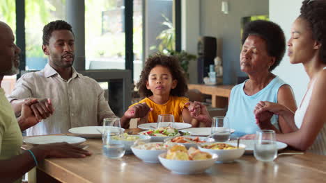 multi-generation family joining hands and saying prayer before meal at home
