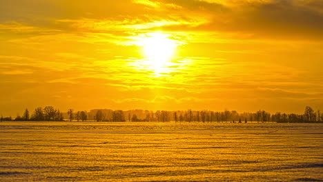 Blick-Auf-Die-Untergehende-Sonne-Im-Zeitraffer-Am-Abend-über-Schneebedeckte-Landwirtschaftliche-Felder-Mit-Autos,-Die-Sich-In-Der-Ferne-Entlang-Der-Autobahn-Bewegen