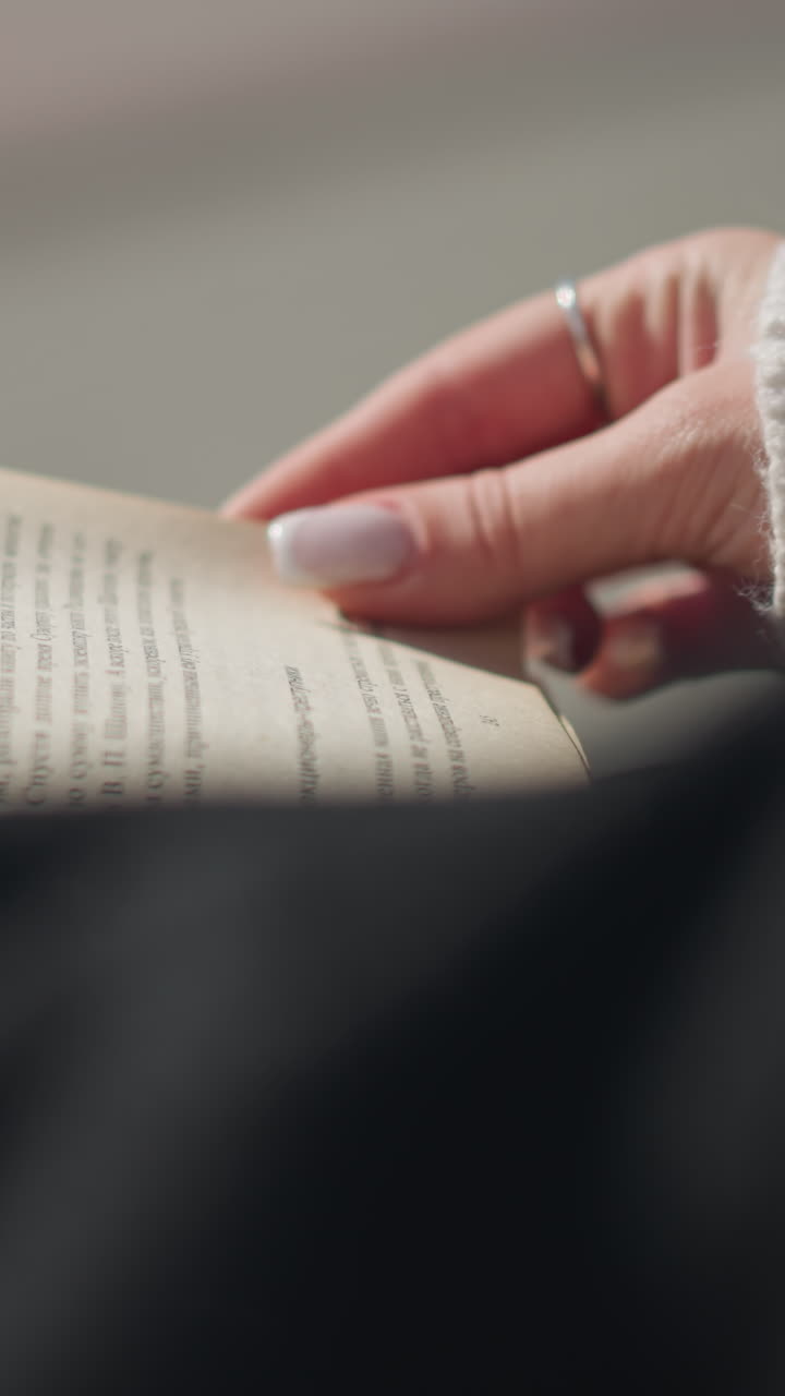 Close-up of a woman s hand with manicured nails gently turning pages of a book, with vibrant page edges and sunlight reflecting off her hand, while soft background