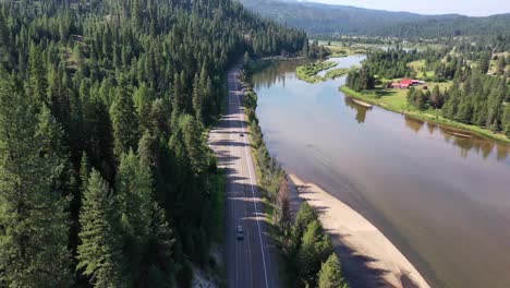 drone pushes in as it passes over a road with busy traffic going both directions. the road is lined with forest trees and runs along a wide river.