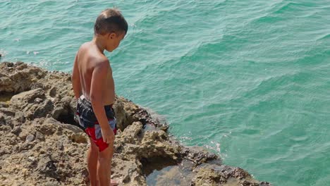 Caucasian-boy-prepares-to-jump-from-rocks-to-the-sea,-Kalamata-,-Greece-,-slow-motion