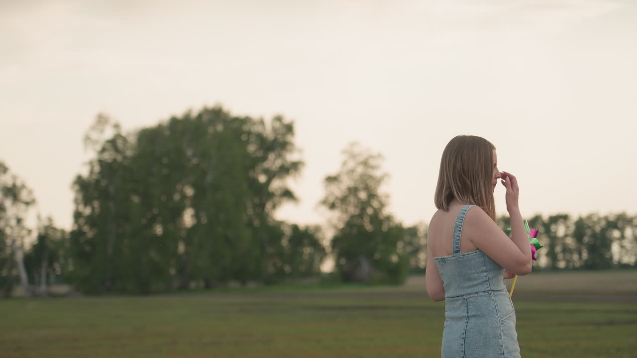 Young woman holding colourful pinwheel looks thoughtfully across vast green field under cloudy summer sky in denim dress capturing pensive mood and gentle breeze