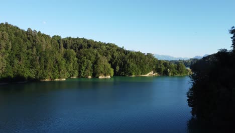 drone-climbs-and-flies-over-the-lac-de-gruyere-in-beautiful-Switzerland,-a-deep-green-little-lake-with-a-lot-of-forest-and-the-white-Alps-in-the-background