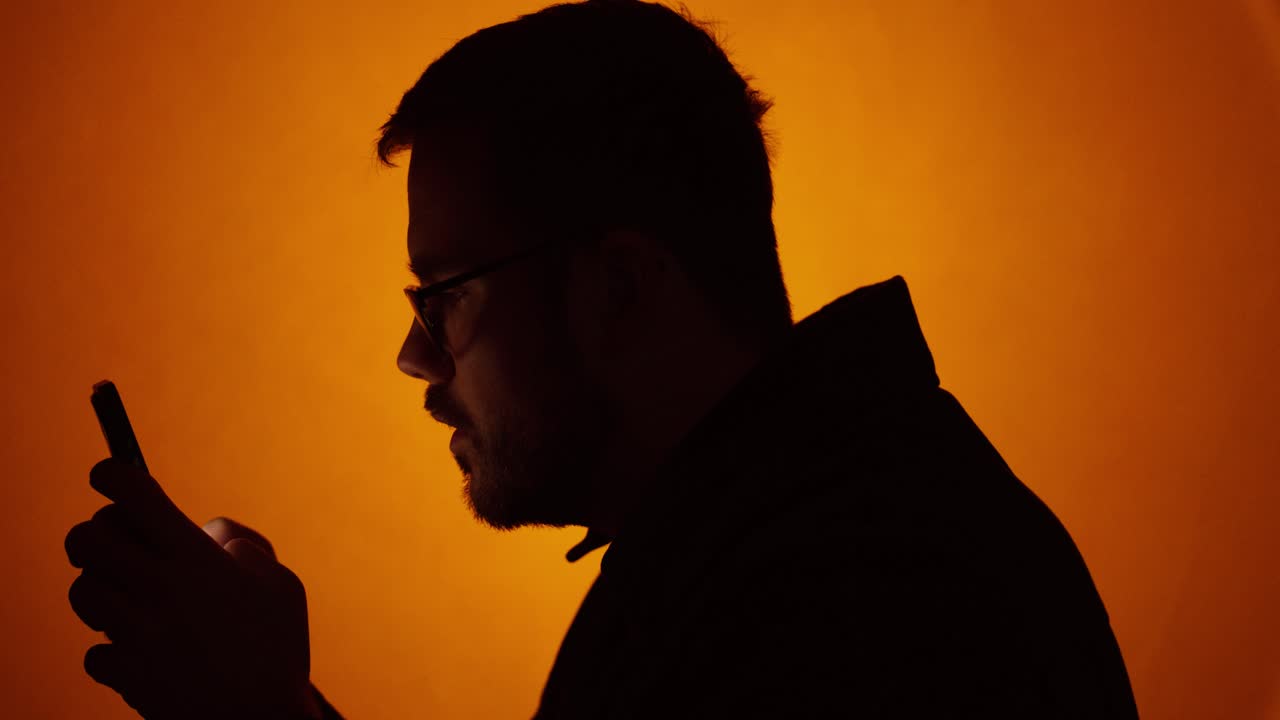 Tight side-profile silhouette of a man focused on his smartphone against a warm orange background, creating a cinematic and anonymous tech mood. Ideal for messaging, digital addiction, privacy concern