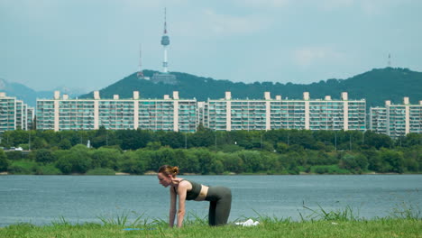 Frau-übt-Oder-Trainiert-Outdoor-Cat-Cow-Stretch-Yoga-Pose-Im-Han-Fluss-Park-In-Seoul-Mit-Ikonischem-Blick-Auf-Den-Namsan-Turm-–-Weitwinkelstatik