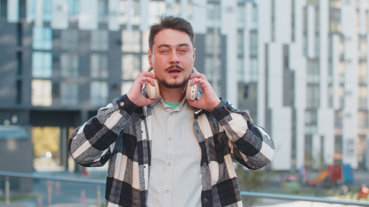Caucasian mature man taking off wireless headphones looking at camera and smiling on city street