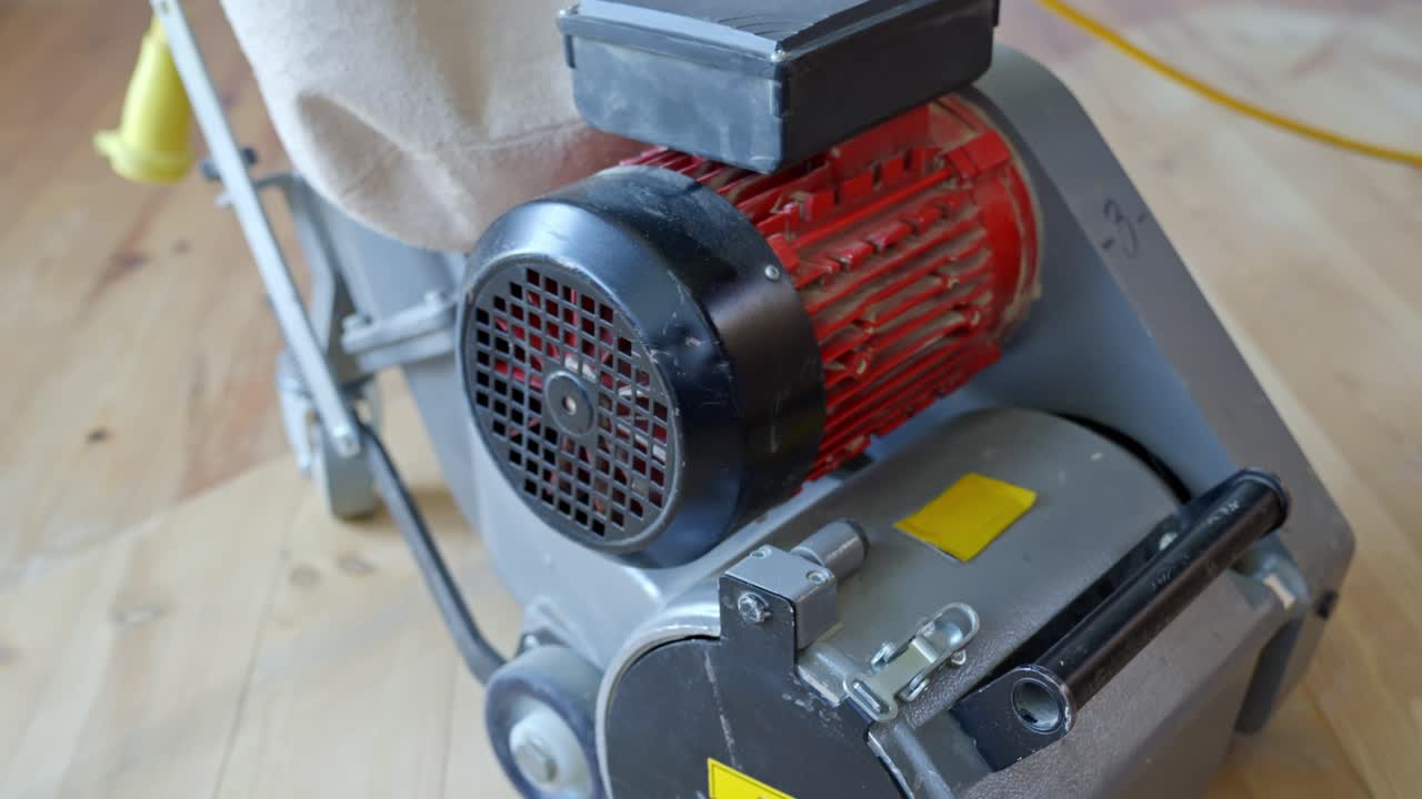 Electric wood floor sanding machine with red and silver motor, dust bag attached, camera pans over the equipment on pine parquet floor during home renovation, highlighting tool components,
