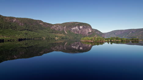 Slow-aerial-shot-approaching-a-small-island-on-Byglandsfjord-in-Norway-on-a-clear-day,-Tree-covered-cliffs-reflect-flawlessly-in-the-still-lake-water