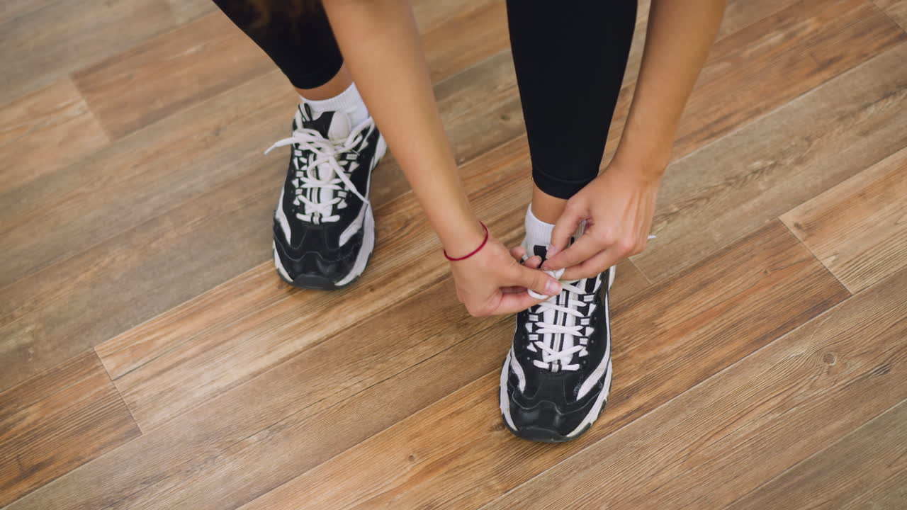 Vista laterale di una donna con leggings neri che si piega in avanti per allacciare le scarpe da ginnastica su un pavimento in legno lucido; le mani tirano forte i lacci, braccialetto rosso visibile sul polso, attenzione focalizzata sulle calzature
