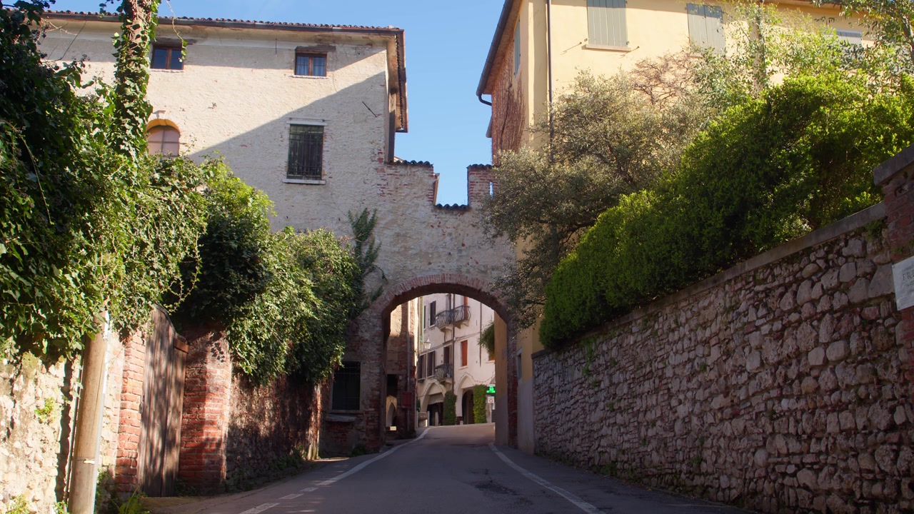 Arco in pietra storico e strada stretta nella città di Asolo illuminata dal sole, incorniciati da vecchie mura.
