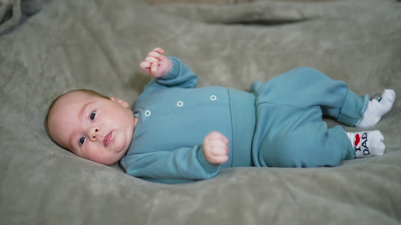 Healthy beautiful toddler lies on the bed and tosses his limbs. Adorable baby boy in warm blue costume on the grey background.