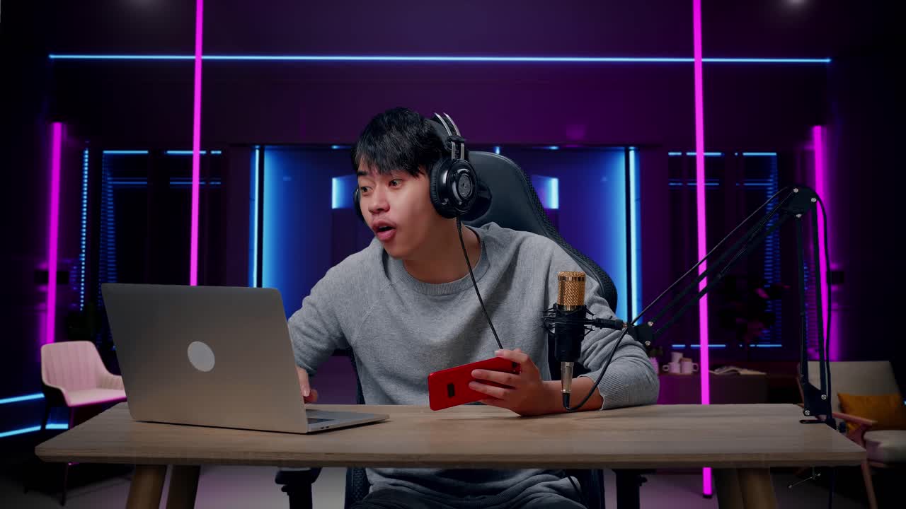 un jugador asiático sonriente con auriculares leyendo comentarios en la computadora mientras usa el teléfono móvil jugando un juego en una habitación iluminada por luces de neón en retro