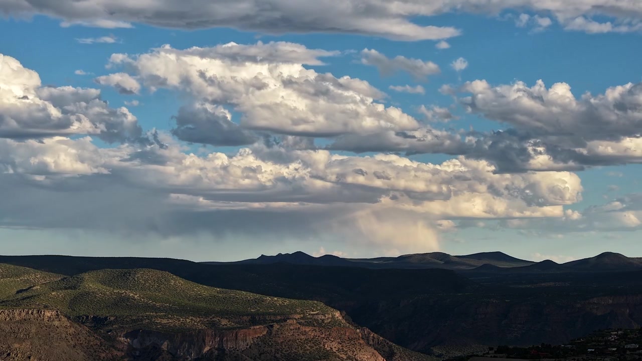 4K UHD drone-timelapse af lagdelte hvide skyer, der bevæger sig hurtigt hen over en blå himmel. Dynamiske bølger af lys og skygge skifter dramatisk over de barske toppe af et frodigt sydvestligt canyonlandskab