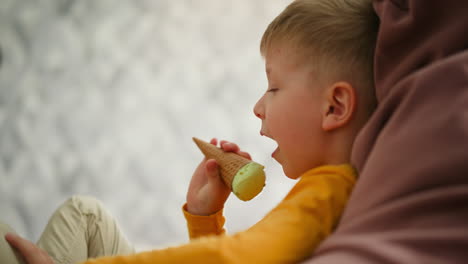 side view of young boy in yellow shirt licking mint chocolate ice cream cone while seated comfortably on pink bean bag with soft blur geometric patterned background