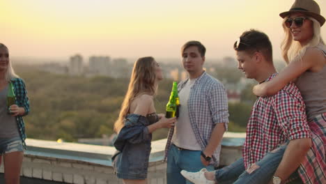 Una-Pareja-Amorosa-Se-Mueve-Bailando-En-Una-Fiesta-Con-Amigos-Que-Beben-Cerveza.-Una-Chica-Sentada-En-La-Espalda-De-Su-Novio-Están-Jugando-En-El-Techo.