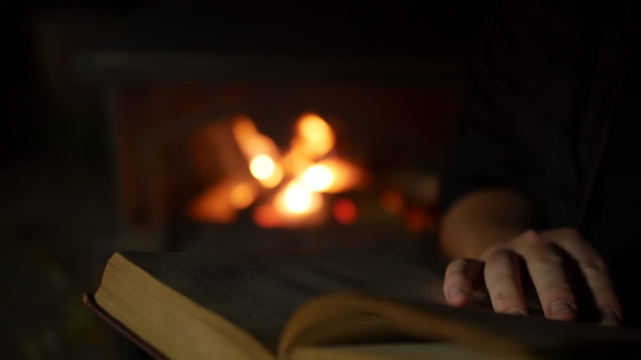 un hombre leyendo y pasando las páginas de un libro cerca de un fuego abierto en una chimenea en una habitación oscura