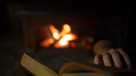 a man reading and turning the pages of a book close to an open fire in a fireplace in a dark room