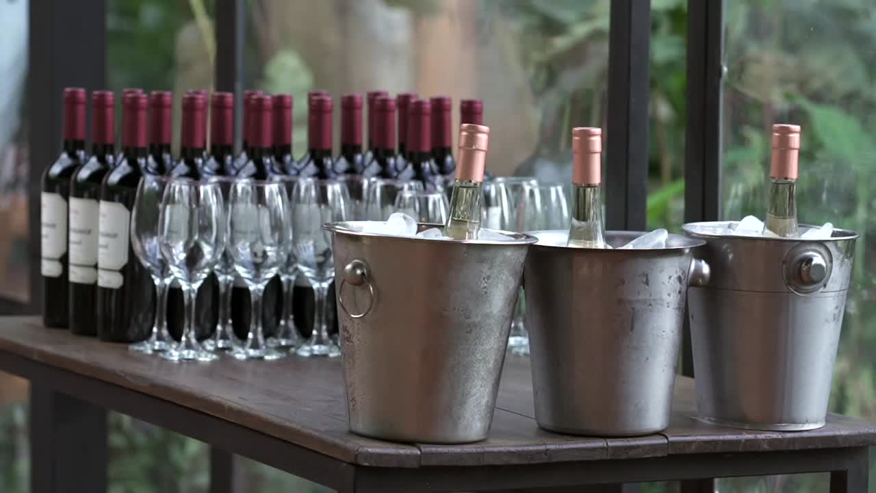 Detailed closeup shot of wooden bar table stocked with red and white wine bottles, glasses, and ice buckets