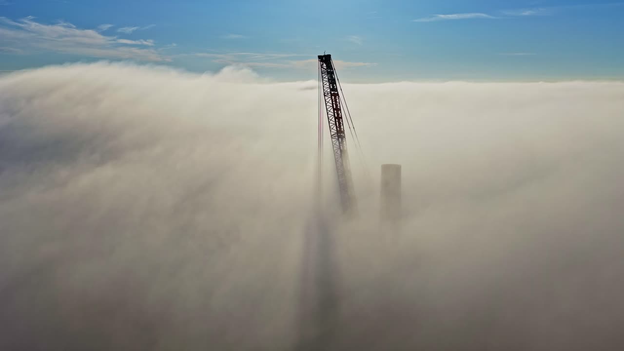 Una gru edile alta trafigge una fitta nebbia mattutina, con la sua silhouette che si erge sopra un soffice strato di nuvole bianche sotto un cielo azzurro limpido