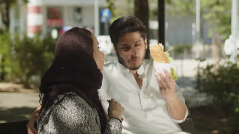 Young-muslim-couple-sitting-on-bench,-eating-snack-on-sunny-day.