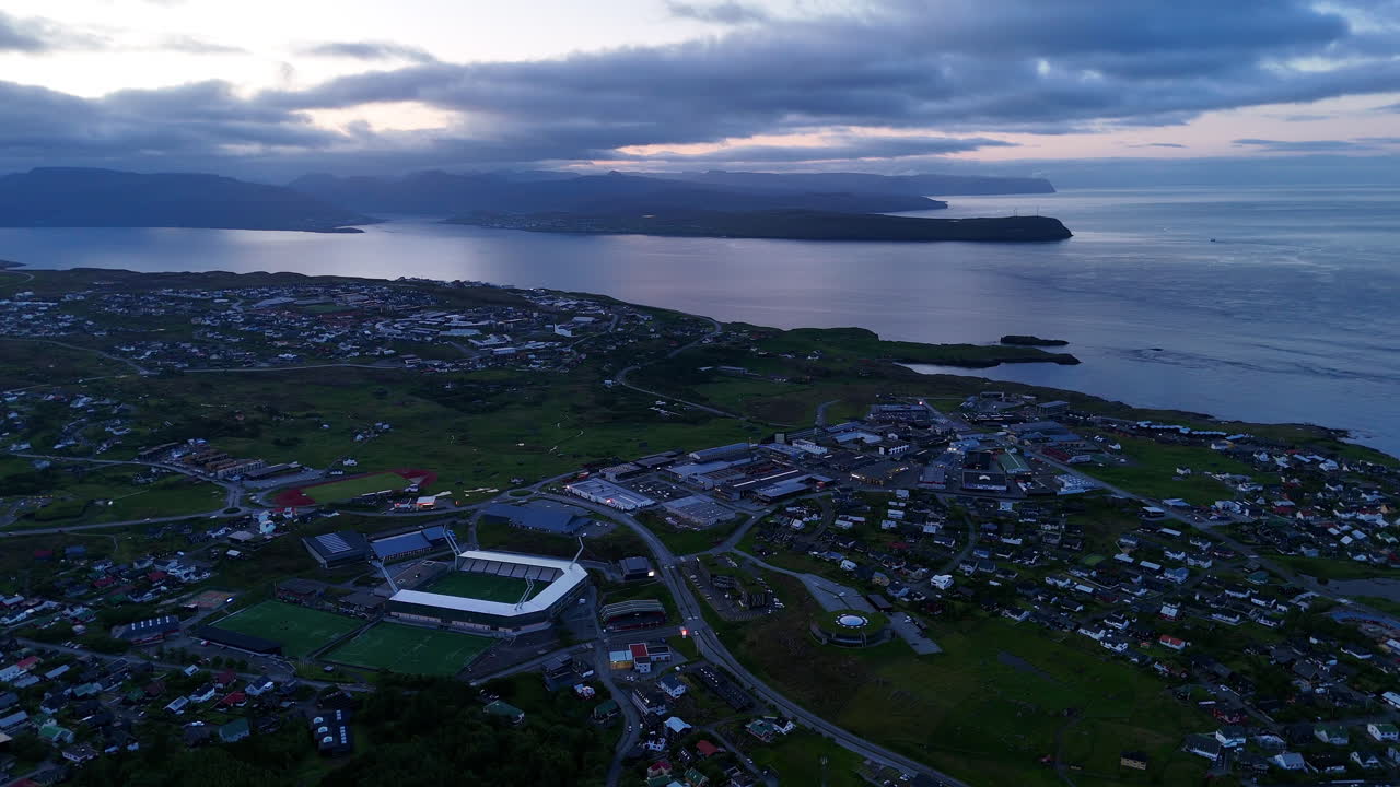 Cinematic aerial view of colorful village houses along a fjord in the Faroe Islands, surrounded by dramatic green mountains, black sand beach, and misty coastal scenery