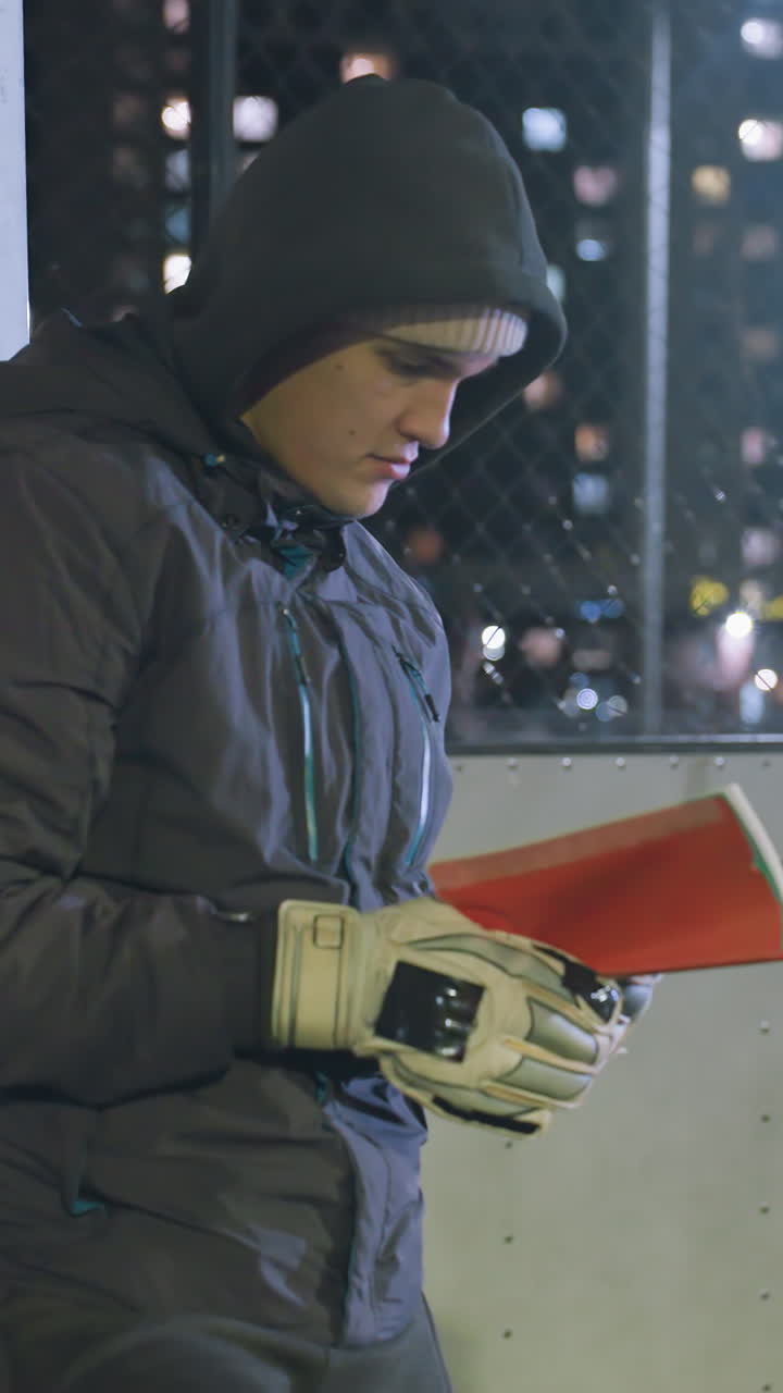 portero con guante de mano en capucha y gorra apoyado contra un poste de meta metálico leyendo un libro al aire libre por la noche, iluminado por las luces de la ciudad