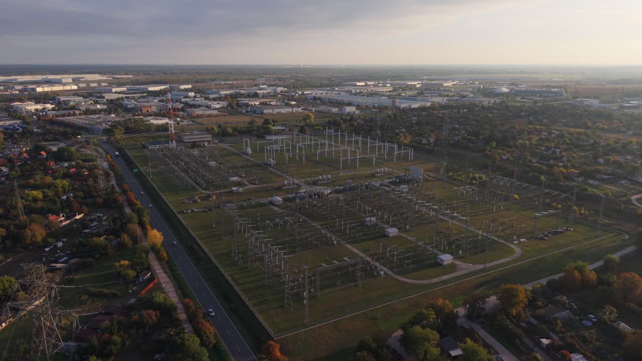 An aerial view of a large electrical substation surrounded by industrial buildings and residential areas. The video is taken during sunset, highlighting the power infrastructure and transmission lines