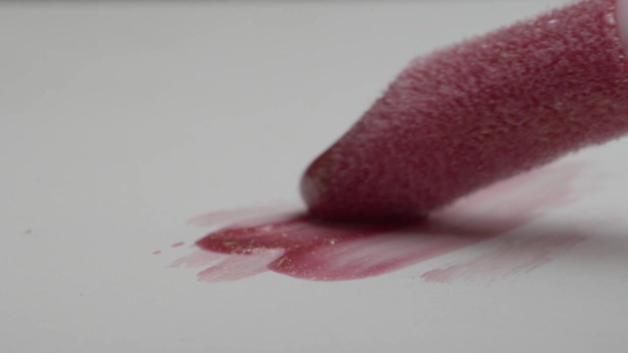 Makeup applicator coated with red cosmetic powder hovering above a white surface. Soft pigment marks and fine applicator texture create an abstract beauty closeup.