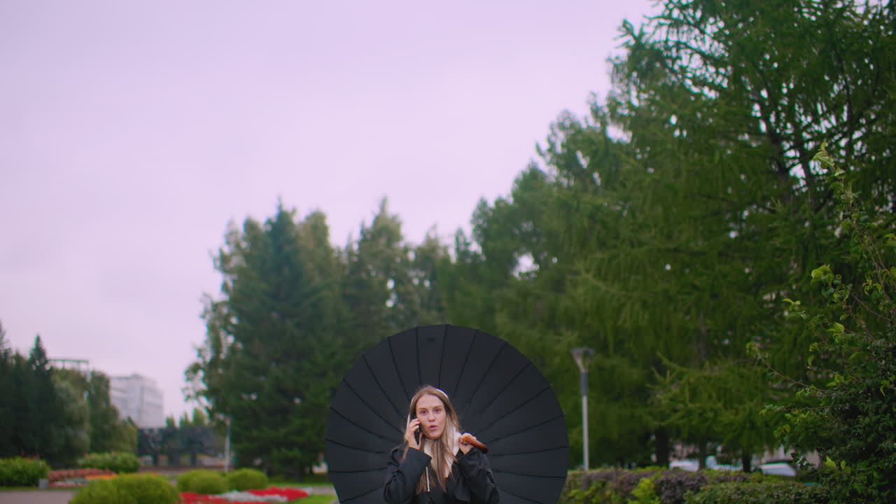 Gorgeous lady in black coat holding umbrella talking on phone in park with green trees and bushes on cloudy rainy day, smiling expression creating relaxed urban outdoor communication lifestyle atmosphere