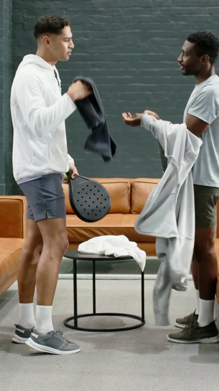 Vídeo vertical: Homem de moletom branco entrando no lounge, segurando uma raquete e uma bolsa de viagem, cumprimentando um amigo para um jogo.