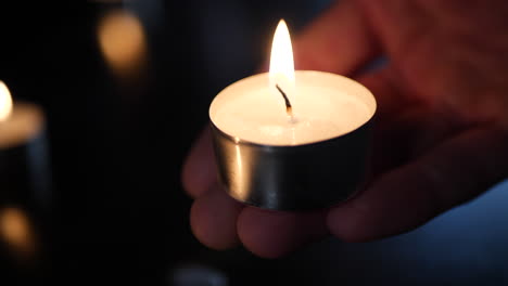 a hand holding a small tealight candle with the flames of many burning in the background during a cande light vigil to mourn the loss of a loved one