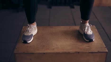 perspective of the bottom part of female sportive workouter jumping up on wooden platform at the gym.