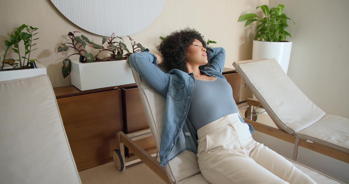 Afroamerikansk voksen kvinde sidder på spa-chaiselong, tager sko af, svinger ben, læner sig tilbage og slapper af