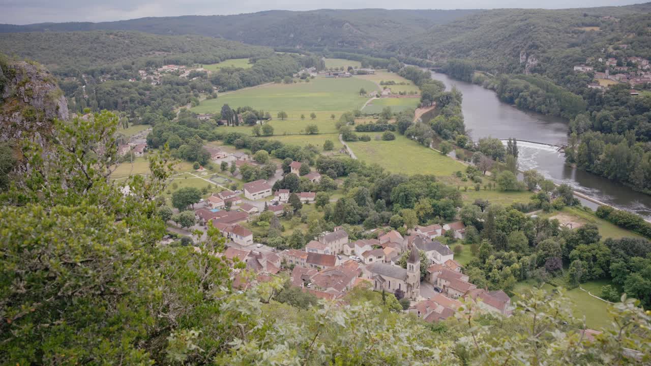 Panorama aereo del villaggio di Saint Géry nella valle del Lot, con l'architettura storica della campagna francese che si erge accanto a un fiume serpeggiante e circondato da verdi colline e scogliere.