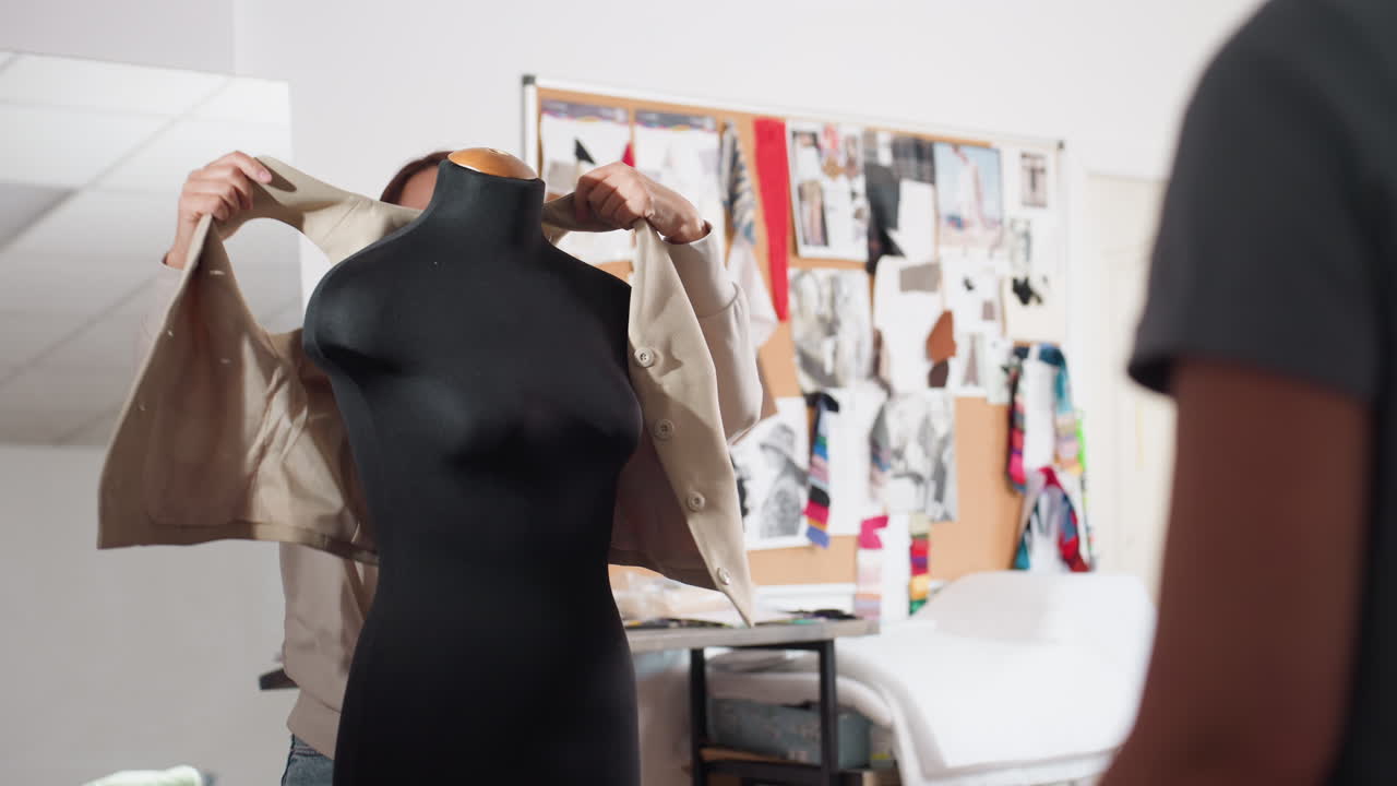 Female designer smiles while sliding beige blazer onto black dress form, chatting with coworker inside studio lined with fabric swatches, sketch filled corkboard, adjusting garment to check fit