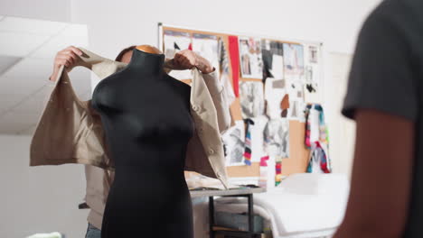 female designer smiles while sliding beige blazer onto black dress form, chatting with coworker inside studio lined with fabric swatches, sketch filled corkboard, adjusting garment to check fit