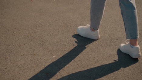 side view of individual wearing jeans and white sneakers walking on tarred road as sunlight casts long shadow beside them, captures casual movement