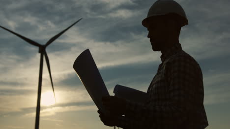 Silhouetten-Einer-Kaukasischen-Frau-Und-Eines-Ingenieurs,-Die-Einen-Helm-Tragen-Und-Einige-Dokumente-Oder-Blaupausen-An-Einer-Windstation-Für-Erneuerbare-Energien-Lesen