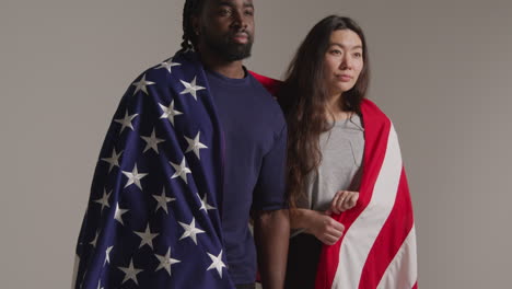 studio portrait shot of multi-cultural couple wrapped in american flag celebrating 4th july independence day