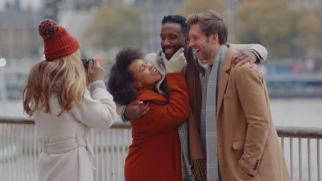 group of friends outdoors wearing coats and scarves posing for photos on camera on autumn or winter trip to london - shot in slow motion