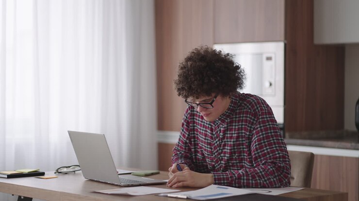 En attraktiv, krølhåret mandlig studerende med briller studerer hjemme. Han bruger sin bærbare computer til at skrive og tage noter i en notesbog. Den studerende ser et online fjernundervisningsseminar på sin bærbare computer.