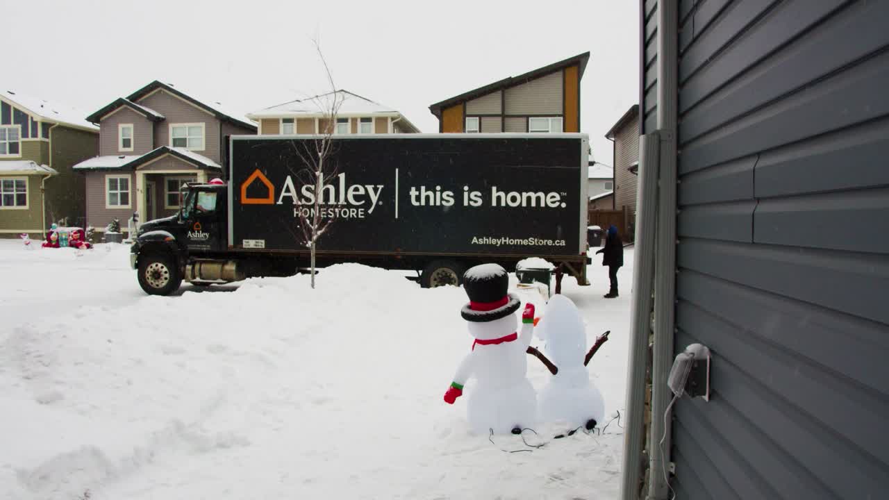 Winter shot of Ashley HomeStore truck at end of snowy lane delivery workers unloading, neighborhood houses in background