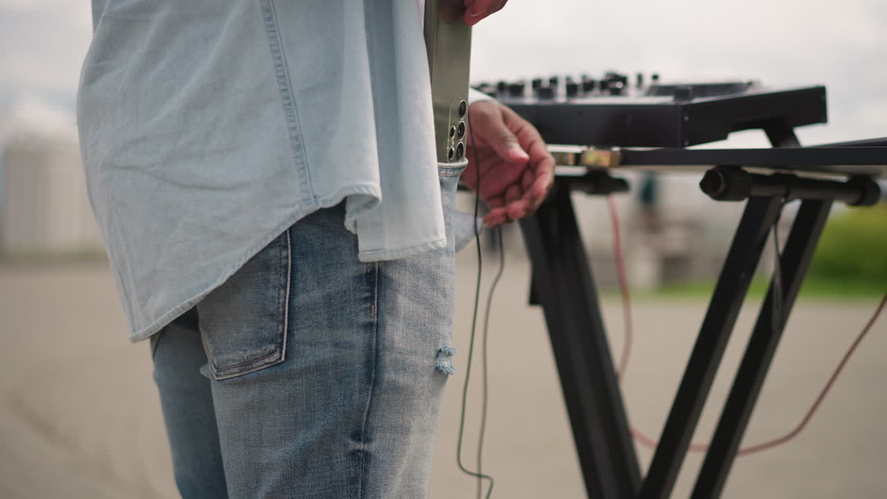 DJ di strada all'aperto che allestisce un mixer su pavimentazione urbana, primo piano di una mano che tiene cuffie e cavi accanto a un controller compatto, abbigliamento casual in denim, cielo coperto, umore di attesa che si prepara per l'aperto.