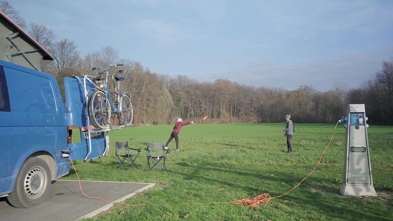 En mand og kvinde kaster frisbee mellem sig med en blå autocamper i forgrunden. De leger på en campingplads' mark med strøm tilsluttet autocamperen i Slovenien