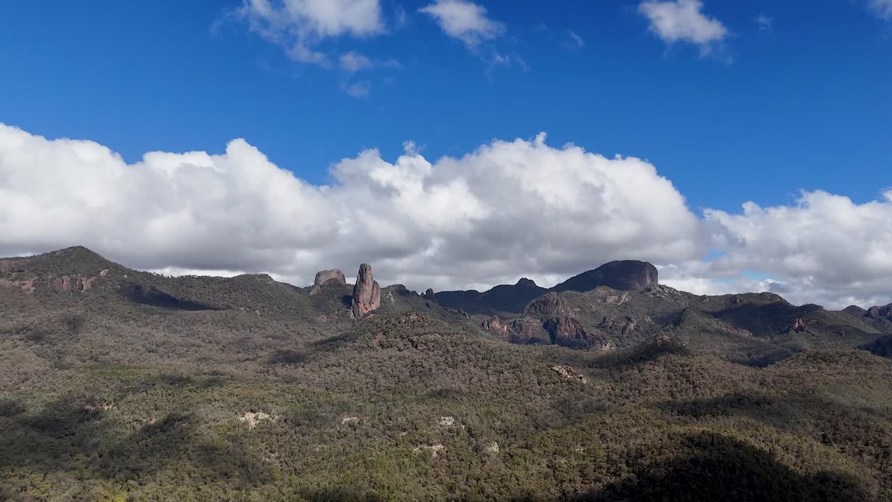 Drone camera smoothly pans across rugged volcanic peaks and forested hills under bright daylight, revealing dramatic clouds and expansive Australian wilderness