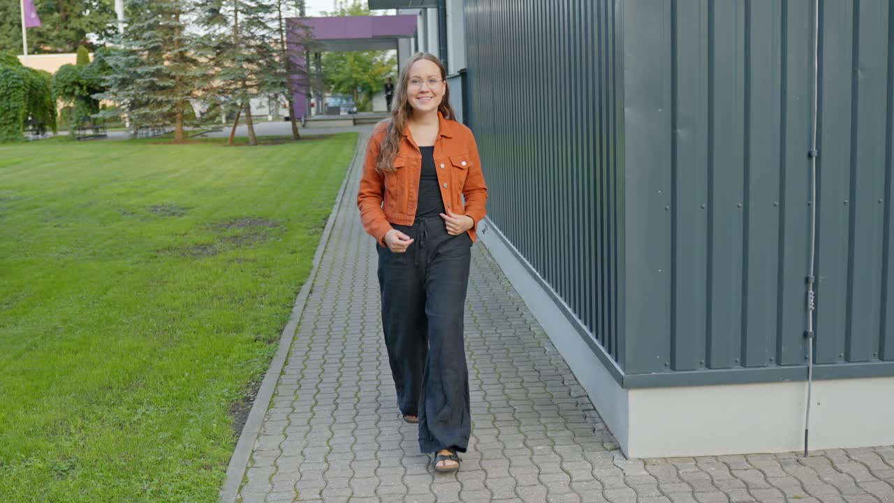 Verspielte, süße Brünette in schwarzem Pullover und orangefarbener Jacke, die freudig lächelnd durch eine urbane Umgebung geht.