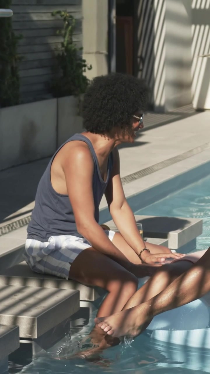 Vertical video: Turning man in shades, shorts, talking and splashing at pool edge, light shifting