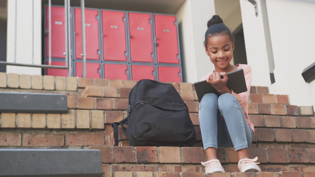 Sitting on school steps, African American girl using tablet and smiling with backpack nearby, copy s