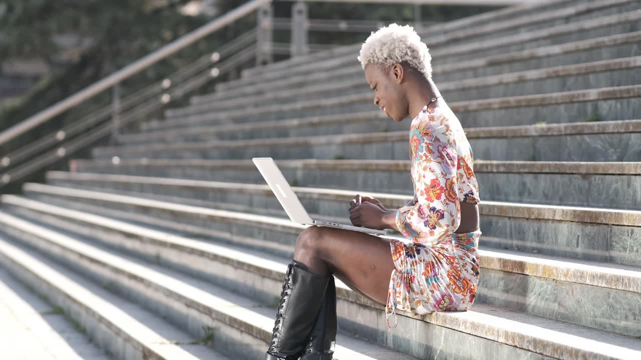 Positive black transgender person typing on laptop on stairs