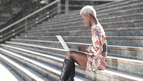 positive black transgender person typing on laptop on stairs